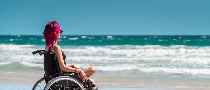 Frau im Rollstuhl mit einem Bein sitzt entspannt am Strand und blickt aufs Meer