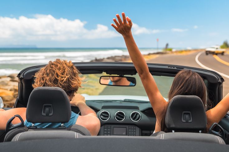 Zwei Personen im Auto mit offenem Verdeck, im Hintergrund kann man das Meer sehen