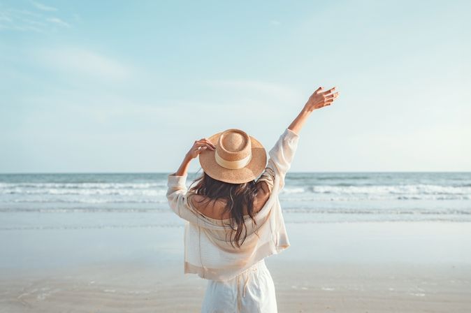 Eine Frau mit Hut von hinten, Sie schaut vom Strand aus auf das Meer und hebt den Arm 