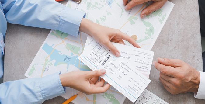 Auf einem Tisch liegen eine Landkarte, ein Taschenrechner, ein Reisepass und eine Geldbörse. Zwei Paar Hände sind zu sehen, ein Paar Hände hält Flugtickets