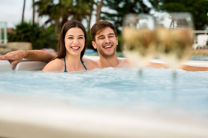Eine Frau und ein Mann sitzen in lachend in einem Whirlpool, verschwommen sieht man im Vordergrund Weingläser