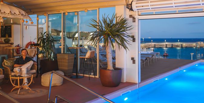 Ein halbüberdachter Pool am Abend, unter der Überdachung stehen Pflanzen und Stühle, auf einem der Stühle sitzt ein Mann