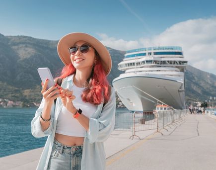 Eine Frau mit roten Haaren steht an einem Hafen, hält ihr Smartphone fest und lächelt; Im Hintergrund steht ein Kreuzfahrtschiff