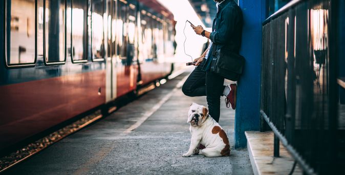 Ein Mann und ein Hund stehen gemeinsam an einem Bahnsteig und schauen in die Kamera