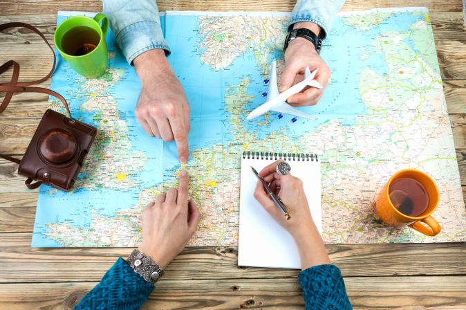 Weltkarte auf Holztisch ausgebreitet, Zwei Personen zeigen mit Fingern auf verschiedene Orte auf der Karte, Eine Person hält ein Modellflugzeug, Notizblock mit Stift, Zwei Tassen stehen auf dem Tisch, Alte Kamera mit Ledertasche