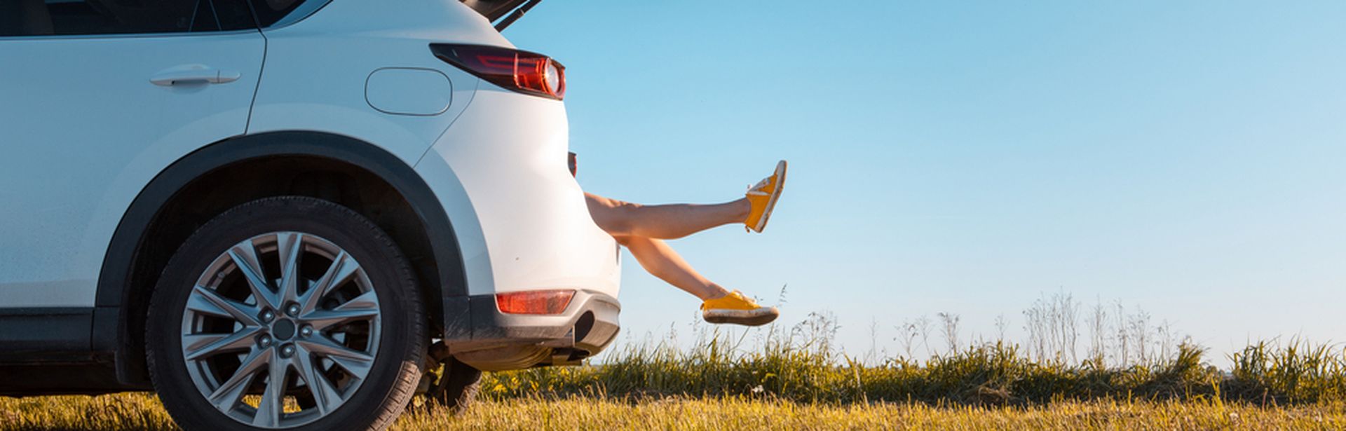 Ein Auto ist zur Hälfte von der Seite zu sehen und steht am Straßenrand an einer Wiese. Der Kofferraum ist auf und man sieht Beine herausragen