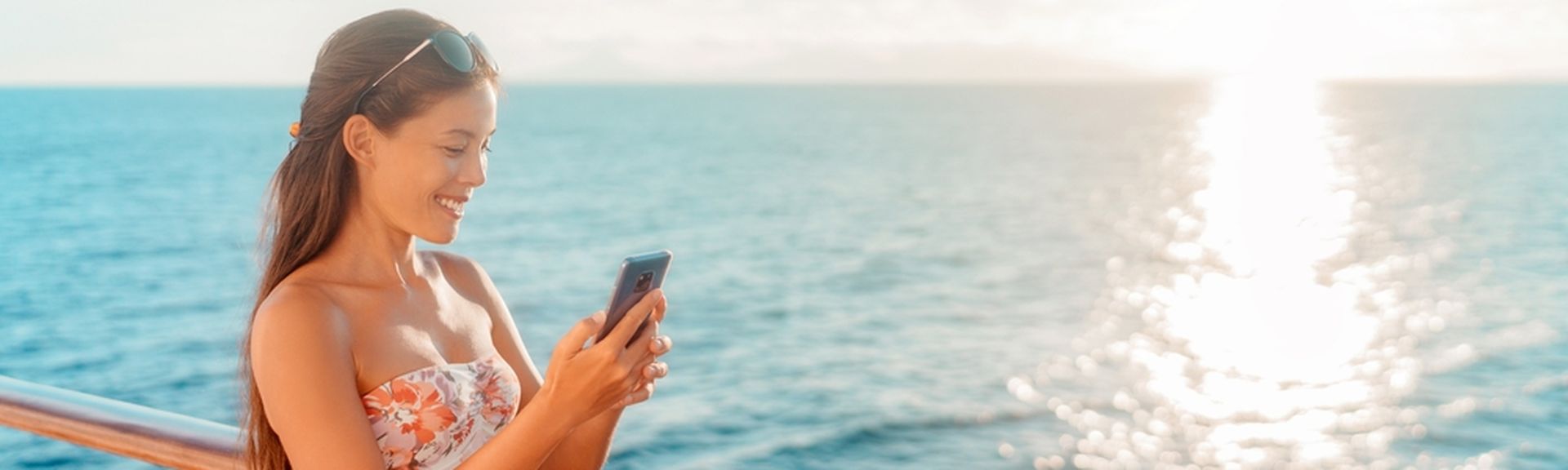 Eine Frau steht lächeln an der Reling eines Kreuzfahrtschiffes und schaut auf ihr Smartphone