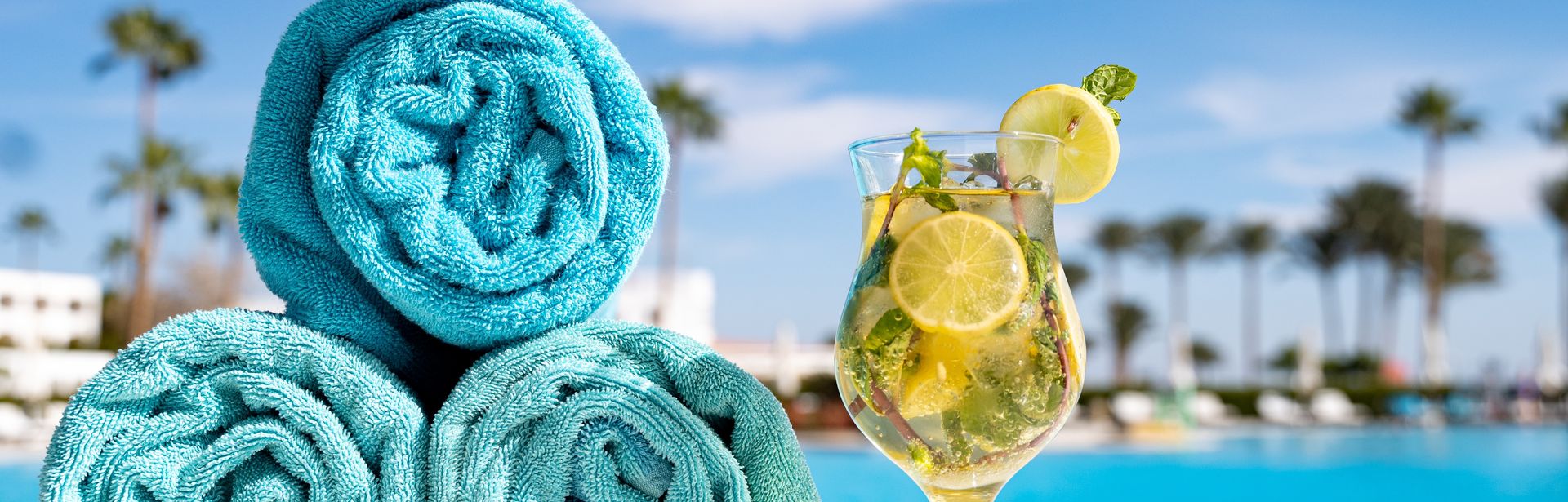 Drei aufgerollte Handtücher und ein Getränk stehen am Pool mit Palmen im Hintergrund