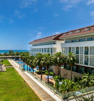 Sentido Trendy Verbena Hotel mit weißer Fassade und ornamentalen Verkleidungselementen, umgeben von Palmen, einem Außenpool mit blauen Sonnenschirmen und gepflegten Grünanlagen – im Hintergrund das türkisblaue Mittelmeer unter strahlend blauem Himmel.