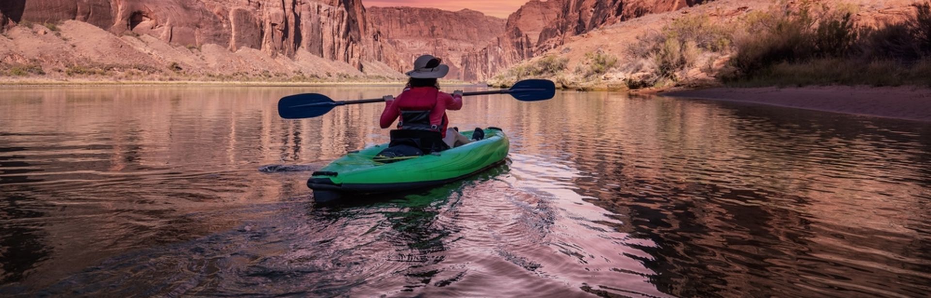 Eine Person sitzt in einem grünen Kanu und schaut auf einen pinken Sonnenauf/untergang