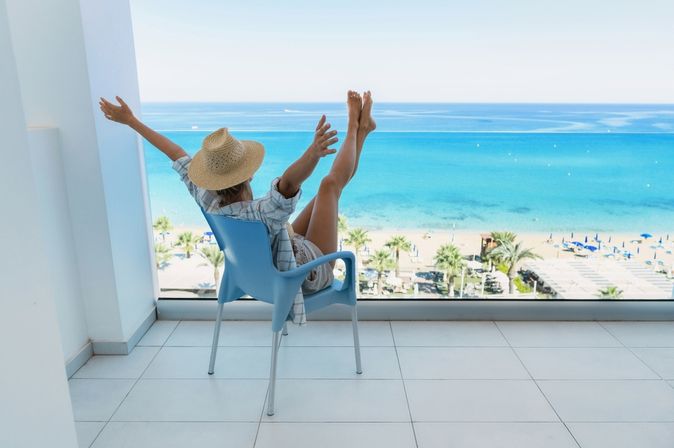 Eine Frau in einem Hemd und einem Strohhut sitzt auf dem Balkon Ihres Hotelzimmers und streckt Arme und Beine in die Luft