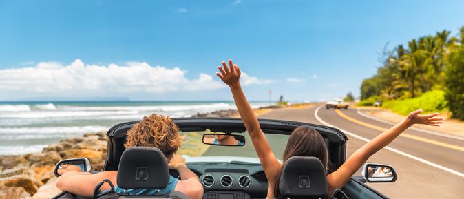 Zwei Personen im Auto mit offenem Verdeck, im Hintergrund kann man das Meer sehen