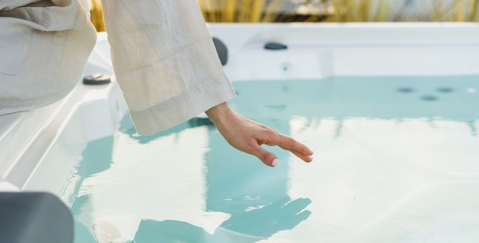 Eine Frau sitzt im Bademantel am Rand eines Whirlpools und hält Ihre Hand über die Wasseroberfläche