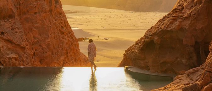 Person läuft am Rand eines Infinity Pools entlang, der zwischen Felsen steht. Im Hintergrund ist Sand und ein Sonnenuntergang zu sehen