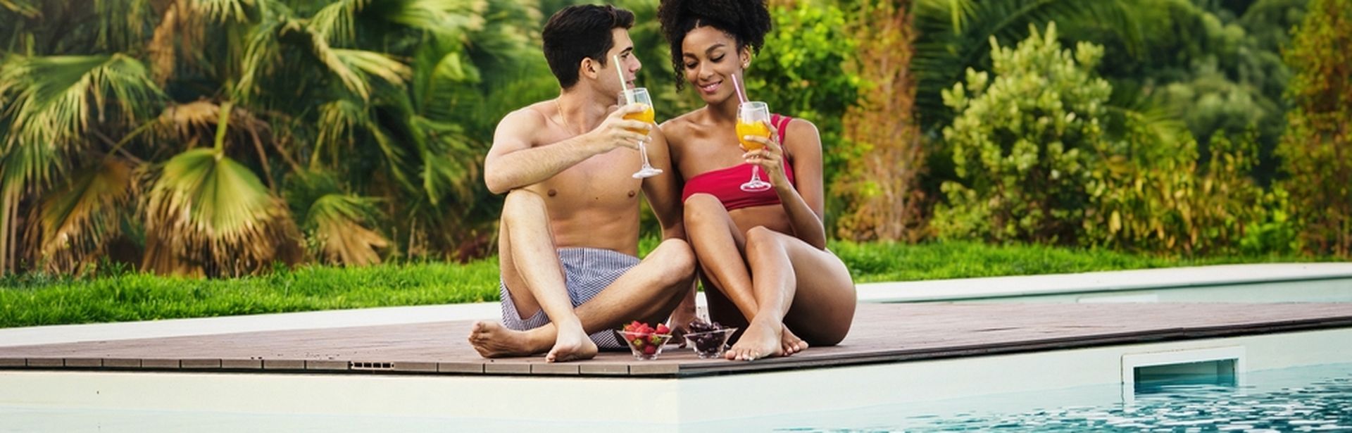 Paar sitzt mit Cocktails in der Hand und Obstschälchen auf der Terrasse vor einem Pool