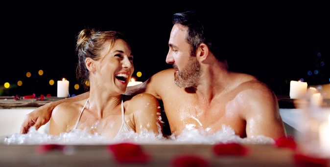 Ein Paar sitzt lachend in einem Whirlpool bei Kerzen und Rosenblättern