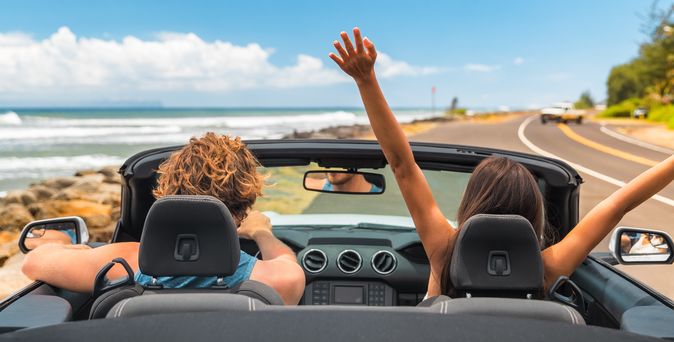 Zwei Personen im Auto mit offenem Verdeck, im Hintergrund kann man das Meer sehen