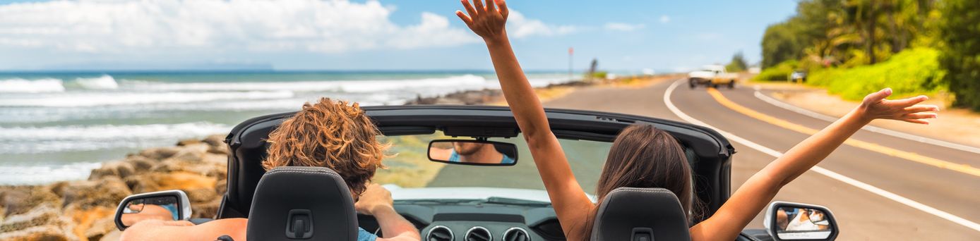 Zwei Personen im Auto mit offenem Verdeck, im Hintergrund kann man das Meer sehen