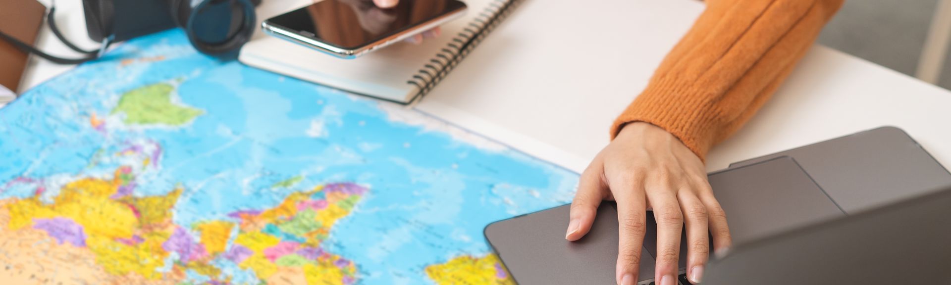 Ein Schreibtisch mit einer Weltkarte als Unterlage, einem Sonnenhut und einer Kamera, eine Person bedient mit der einen Hand ein Handy und mit der anderen einen Laptop