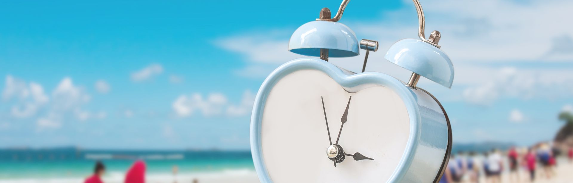 Auf einem Sandhügel steht ein hellblauer, herzförmiger Wecker; Ein Strand mit vielen Menschen ist verschwommen im Hintergrund zu sehen