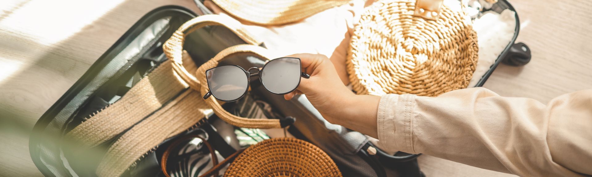 Ein offener Koffer, eine Hand legt eine Sonnenbrille zu den bereits gepackten Dingen