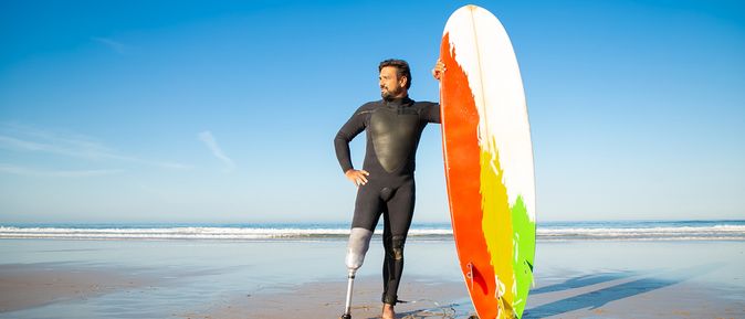 Ein Mann mit einer Beinprothese am linken Bein steht in einem Neoprenanzug am Strand und hält ein buntes Surfboard