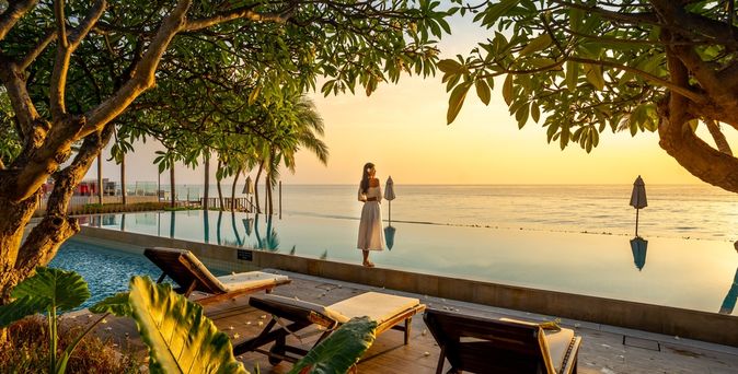 Frau steht bei Sonnenuntergang vor einem Pool der direkt vor einem Meer liegt
