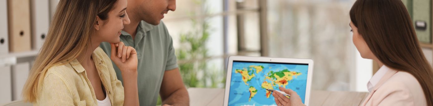 Ein Paar sitzt mit einer Reiseberaterin am Tisch. Die Reiseberaterin zeigt auf eine Weltkarte auf einem Tablet