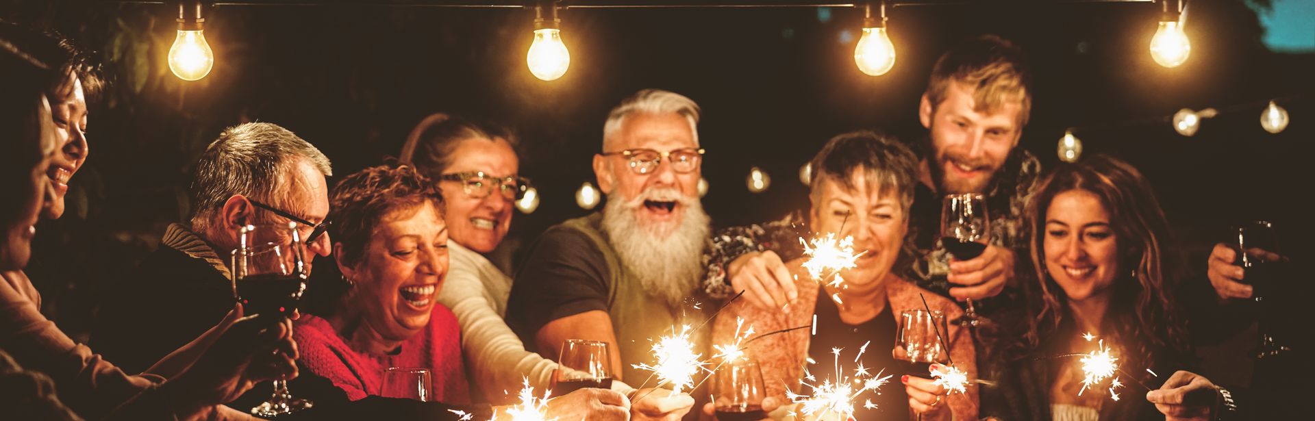 Eine Gruppe von Menschen steht gemeinsam unter einer Lichterkette. Alle halten lachend eine Wunderkerze in die Mitte der Runde.