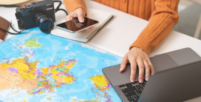 Ein Schreibtisch mit einer Weltkarte als Unterlage, einem Sonnenhut und einer Kamera, eine Person bedient mit der einen Hand ein Handy und mit der anderen einen Laptop