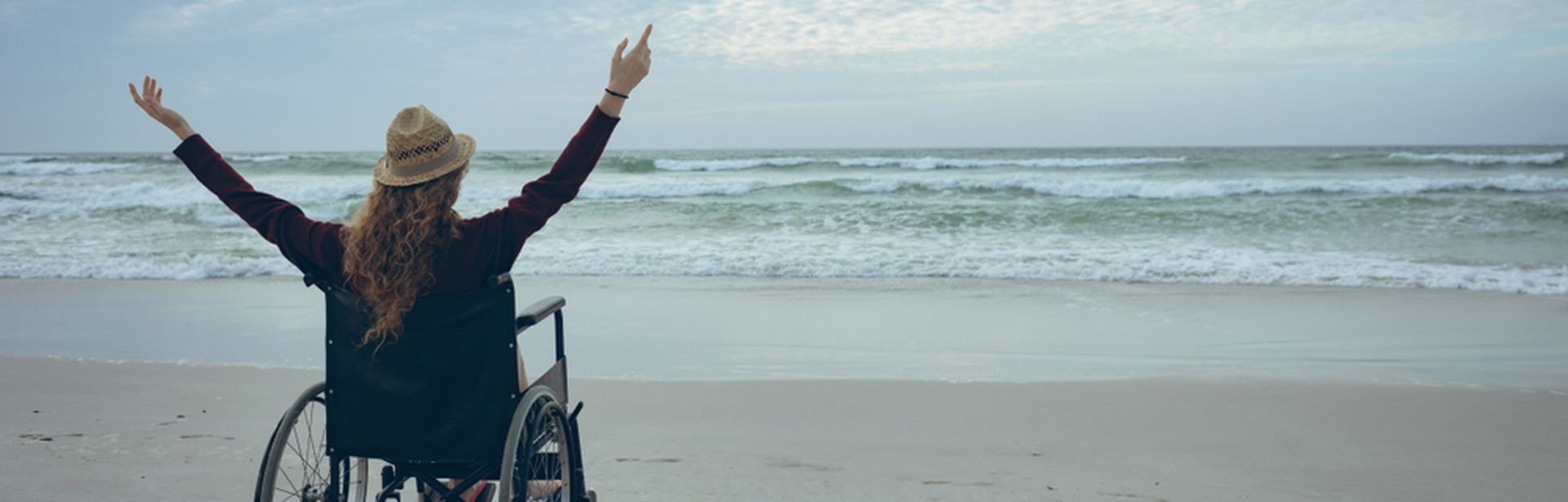 Frau im Rollstuhl hebt die Arme und schaut aufs Meer