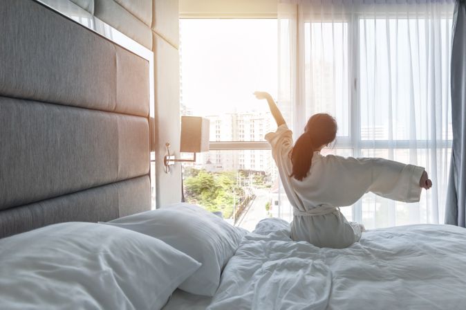 Eine Frau sitzt im Bademantel auf Ihrem Bett und streckt sich 