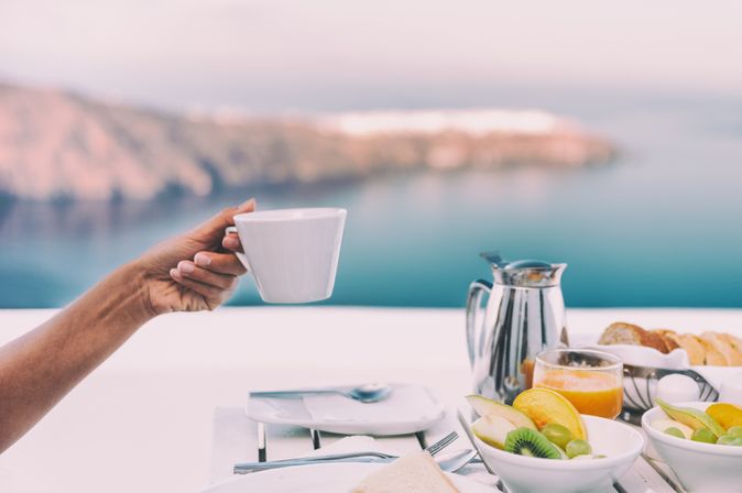 Ein gedeckter Tisch mit einer Kanne und Tellern mit Obst, eine Hand hält eine Tasse hoch, im Hintergrund ist das Meer