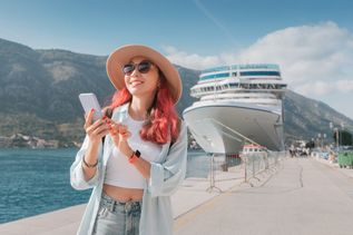Eine Frau mit roten Haaren steht an einem Hafen, hält ihr Smartphone fest und lächelt; Im Hintergrund steht ein Kreuzfahrtschiff