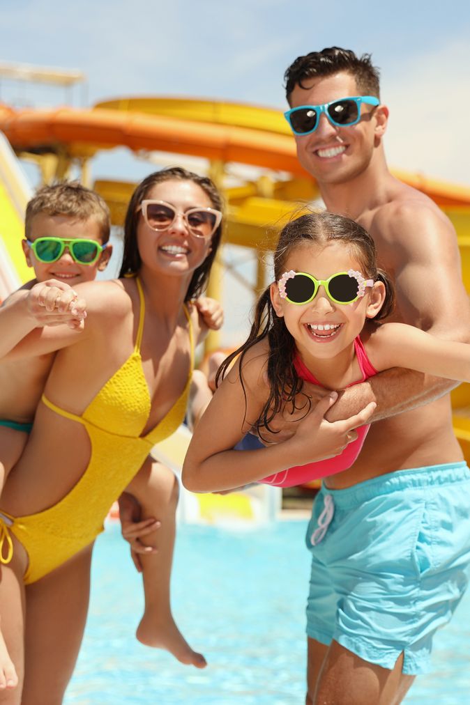 Eine Familie steht in einem Pool vor einigen Wasserrutschen, die Frau hat eins der Kinder auf dem Rücken, der Mann trägt das zweite Kind im Arm 