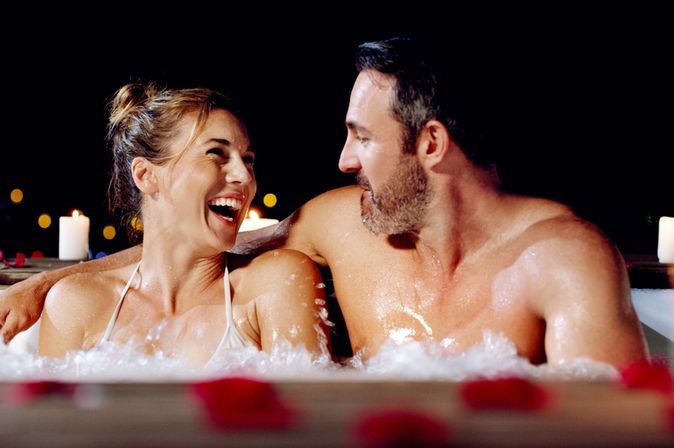 Ein Paar sitzt lachend in einem Whirlpool bei Kerzen und Rosenblättern