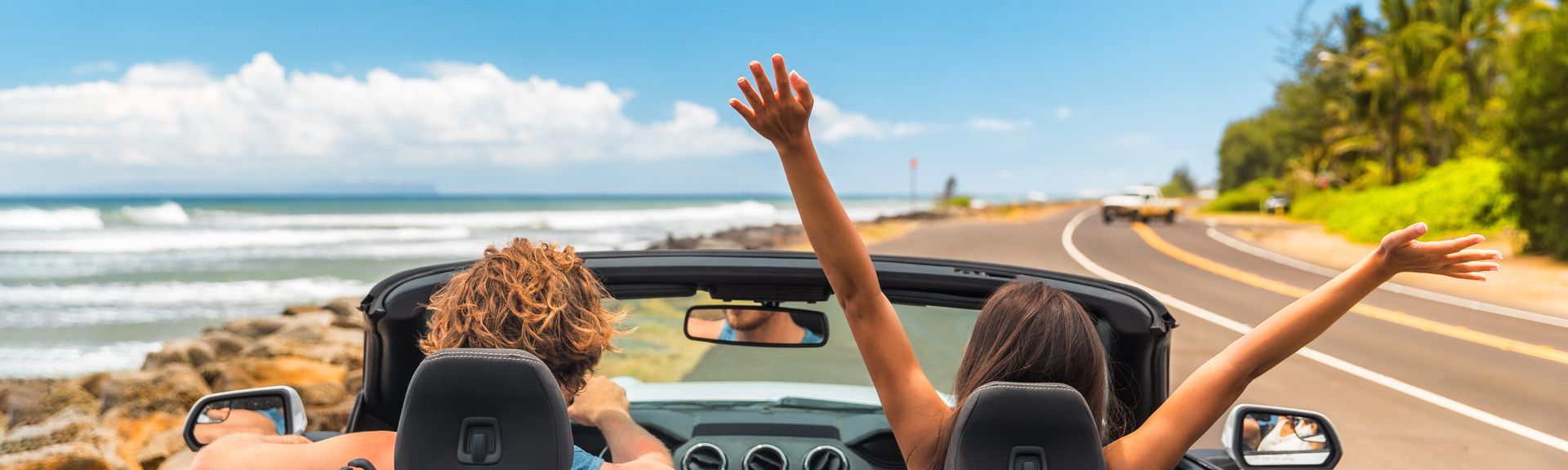 Zwei Personen im Auto mit offenem Verdeck, im Hintergrund kann man das Meer sehen