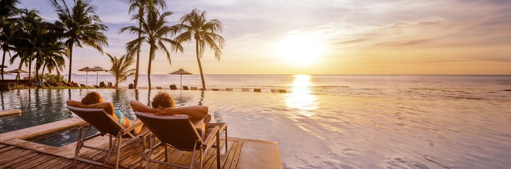 Zwei Personen liegen auf Liegen am Pool und schauen sich den Sonnenuntergang an