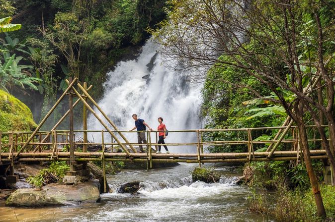 Eine Frau und ein Mann laufen Hand in Hand über eine Holzbrücke über einem Fluss, im Hintergrund ist ein großer Wasserfall