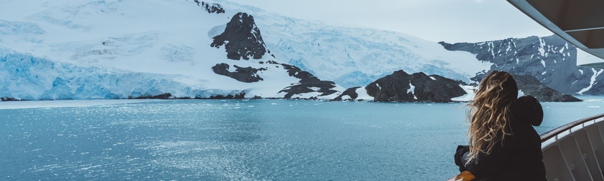 Eine Frau steht mit wehenden Haaren an der Reling eines Kreuzfahrtschiffes und schaut auf schneebedeckte Berge