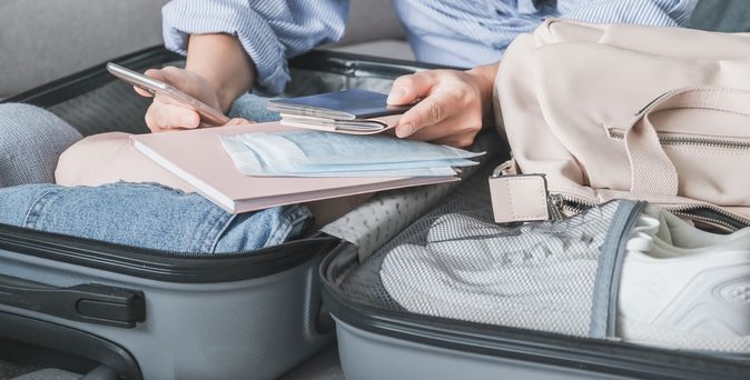 Ein offener Koffer liegt auf dem Sofa. Es ist Kleidung und ein Paar Schuhe im Koffer zu sehen. Eine Hand hält über dem Koffer ein Handy und einige Reiseunterlagen