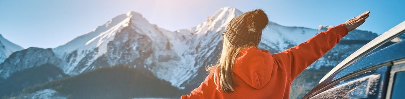 Frau steht an der offenen Tür eines Autos, blickt auf eine schneebedeckte Berglandschaft und streckt die Arme aus