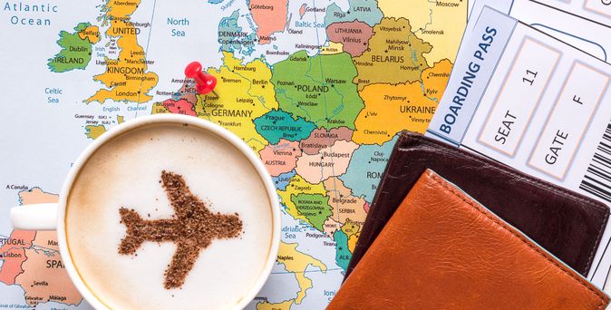 Landkarte mit einem Pin, der in Deutschland gesteckt ist. Auf der Karte steht eine Tasse Kaffee, wo der Schaum die Form einen Flugzeugs ausspart. Ein Flugticket und Lederbörse liegen auch auf der Karte