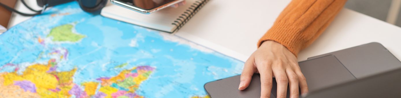 Ein Schreibtisch mit einer Weltkarte als Unterlage, einem Sonnenhut und einer Kamera, eine Person bedient mit der einen Hand ein Handy und mit der anderen einen Laptop