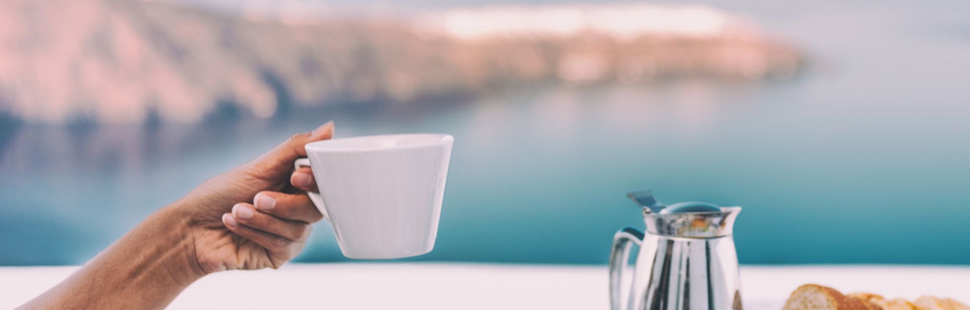Ein gedeckter Tisch mit einer Kanne und Tellern mit Obst, eine Hand hält eine Tasse hoch, im Hintergrund ist das Meer