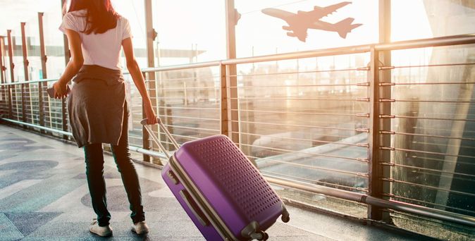 Eine Frau zieht ihren Koffer am Flughafen hinter sich her, draußen hebt gerade ein Flugzeug ab
