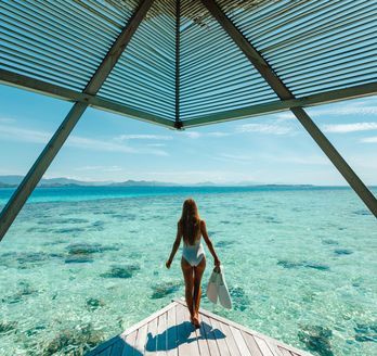 Eine Frau im Bikini steht am Ende eines Stegs und schaut aufs Meer