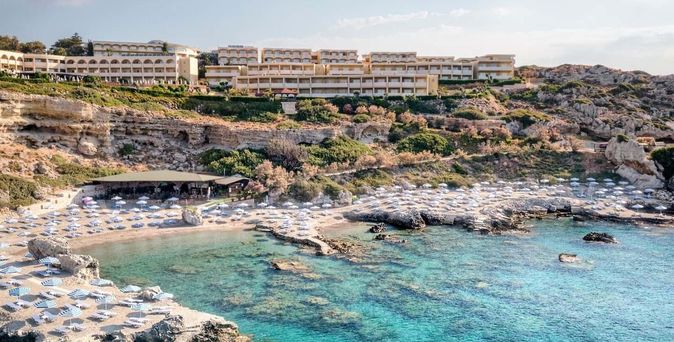 16:11Weitläufige Hotelanlage mit ockergelber Fassade auf einem Felsplateau über einer malerischen Bucht mit türkisklarem Wasser, felsigem Sandstrand und dicht gereihten weißen Sonnenschirmen sowie einem Strandrestaurant.