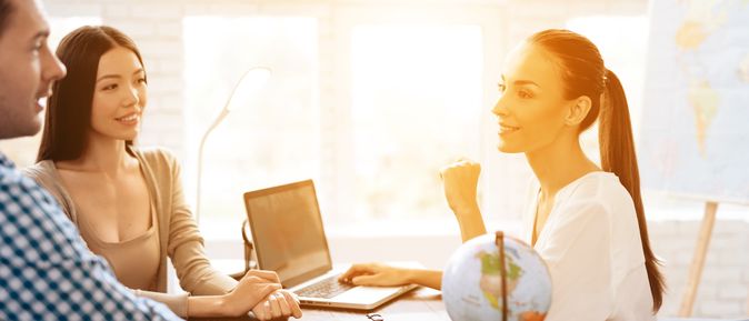 Ein Paar sitzt mit einer Reiseberaterin am Tisch, während das Licht durch das Fenster im Hintergrund den Raum erleuchtet