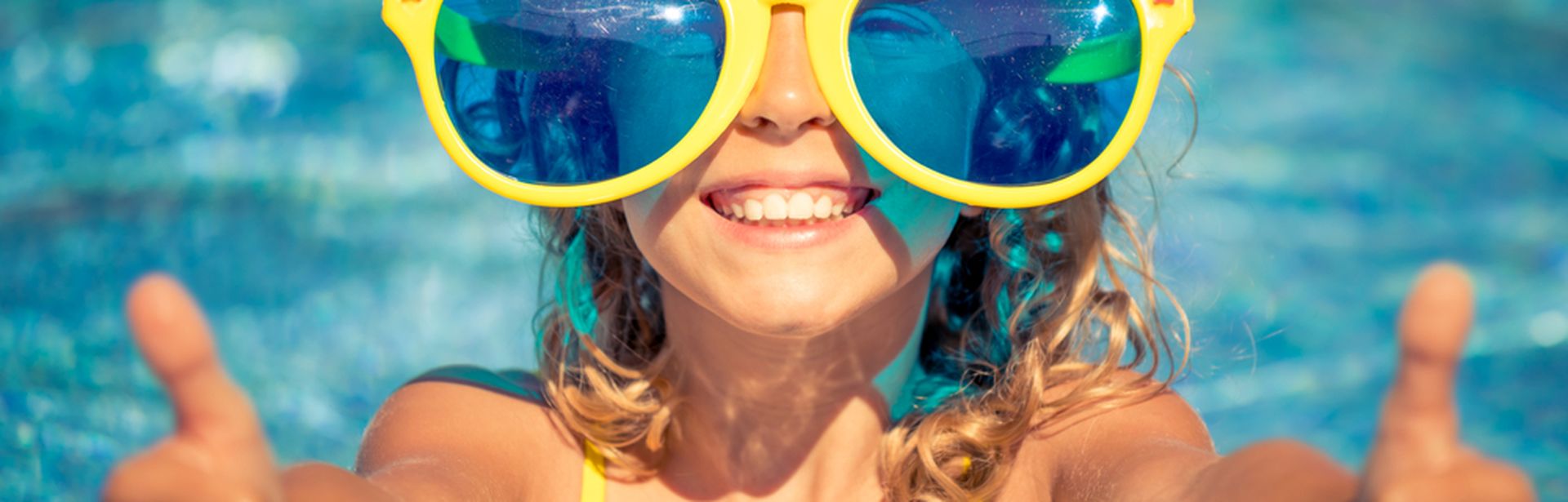 Kind mit Sonnenhut übergroßer Sonnenbrille lächelt und hält beide Daumen nach oben in die Kamera